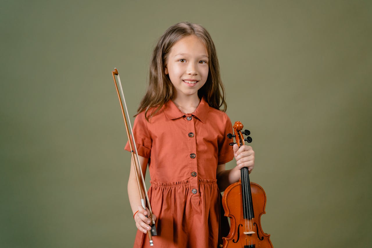 A young girl smiling while holding a violin and bow in a studio setting.