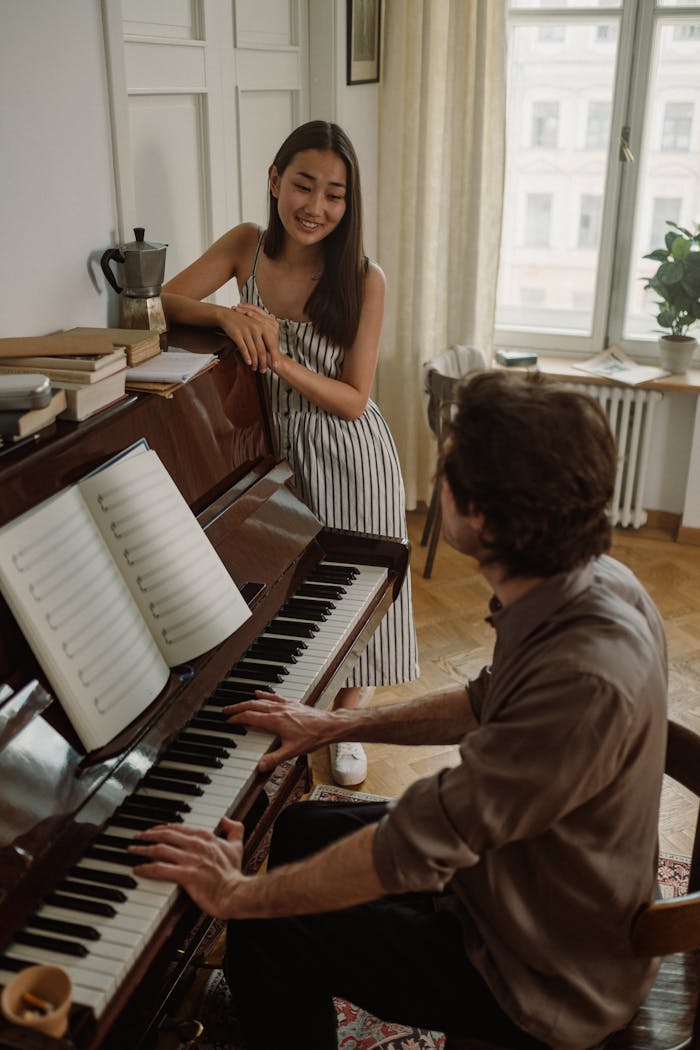 A young woman enjoys listening to a man playing the piano in a cozy indoor setting.